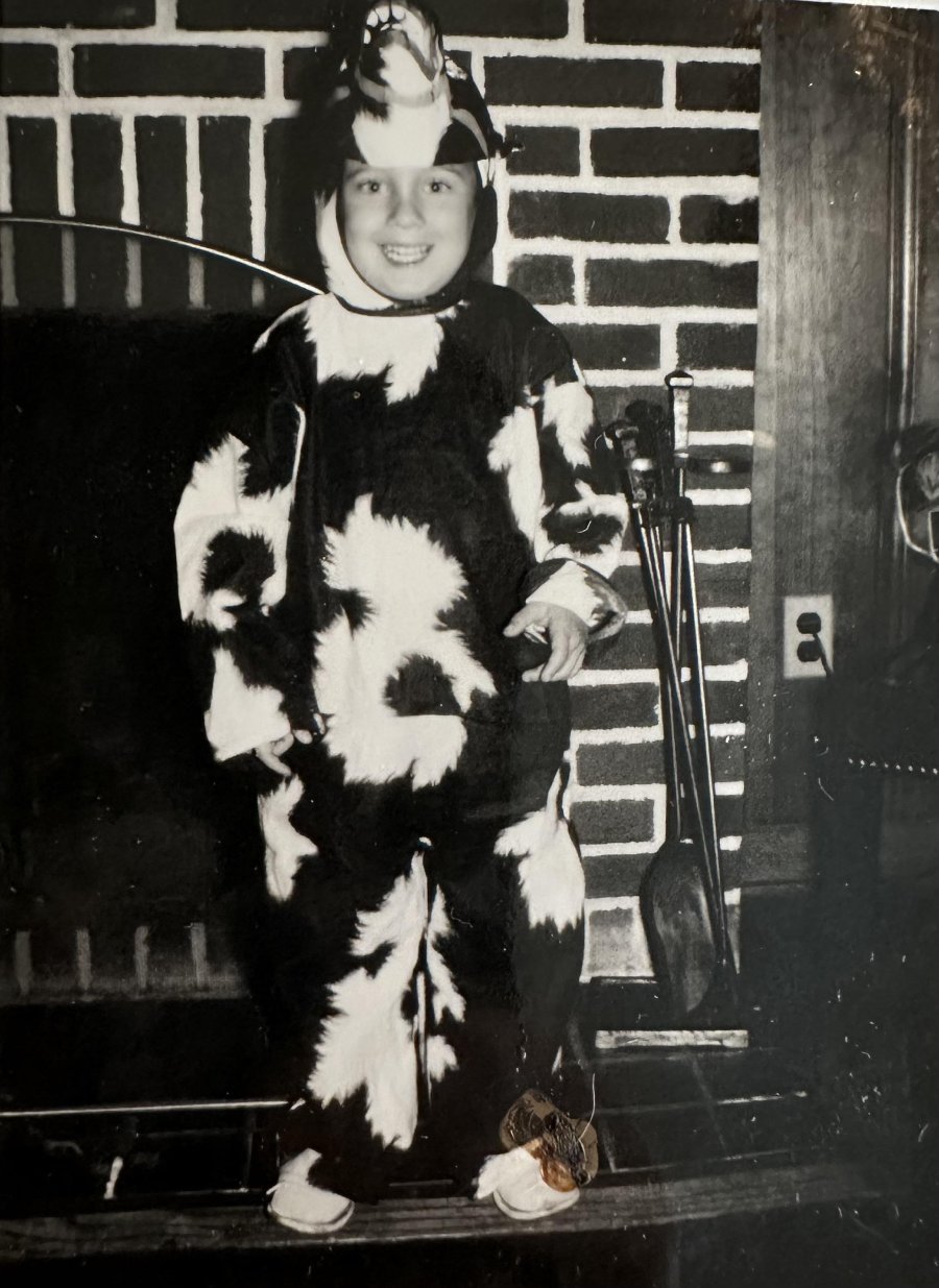 A smiling child in a cow costume stands in front of a brick fireplace, with fireplace tools and a chair visible nearby. The photo is black and white.