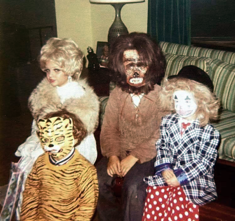 Four children in Halloween costumes sit indoors. Outfits include a blonde wig and fur stole, a werewolf mask, a tiger mask with a striped shirt, and a clown costume with a painted face, hat, and polka-dotted pants.