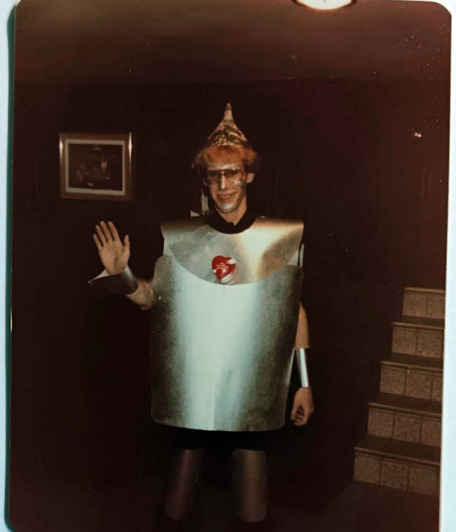 A person wearing a homemade robot costume with a shiny chest plate, a heart sticker, glasses, and a pointed hat waves and smiles while standing indoors near stairs.