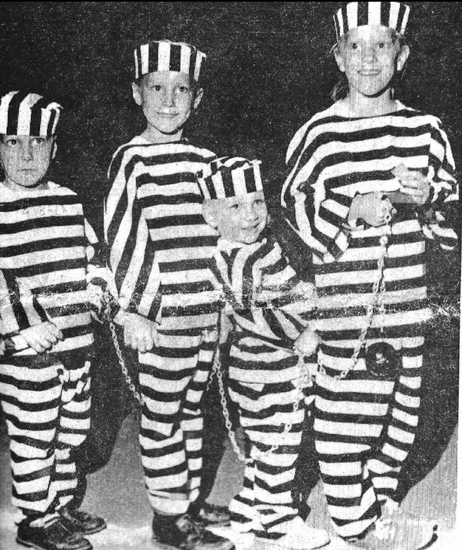 Four children dressed in old-fashioned black and white striped prisoner costumes with hats stand in a line, holding a chain, with one child attached to a ball and chain.