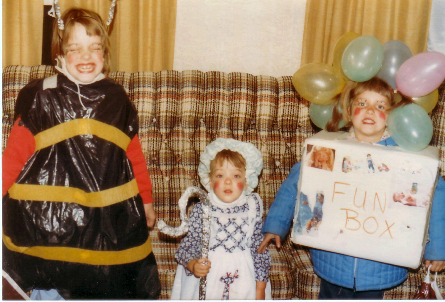 Three children in costumes stand on a plaid couch: one dressed as a bee, one as Little Bo Peep, and one with balloons and a box labeled “Fun Box,” all with face paint and smiling.