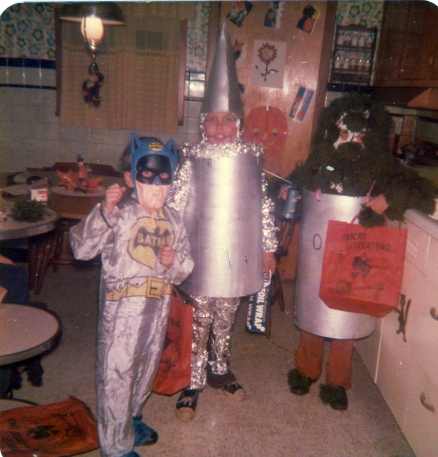 Three children in homemade costumes—Batman, the Tin Man, and a fuzzy green creature—pose in a vintage kitchen holding orange trick-or-treat bags, ready for Halloween. The room has wood cabinets and patterned wallpaper.