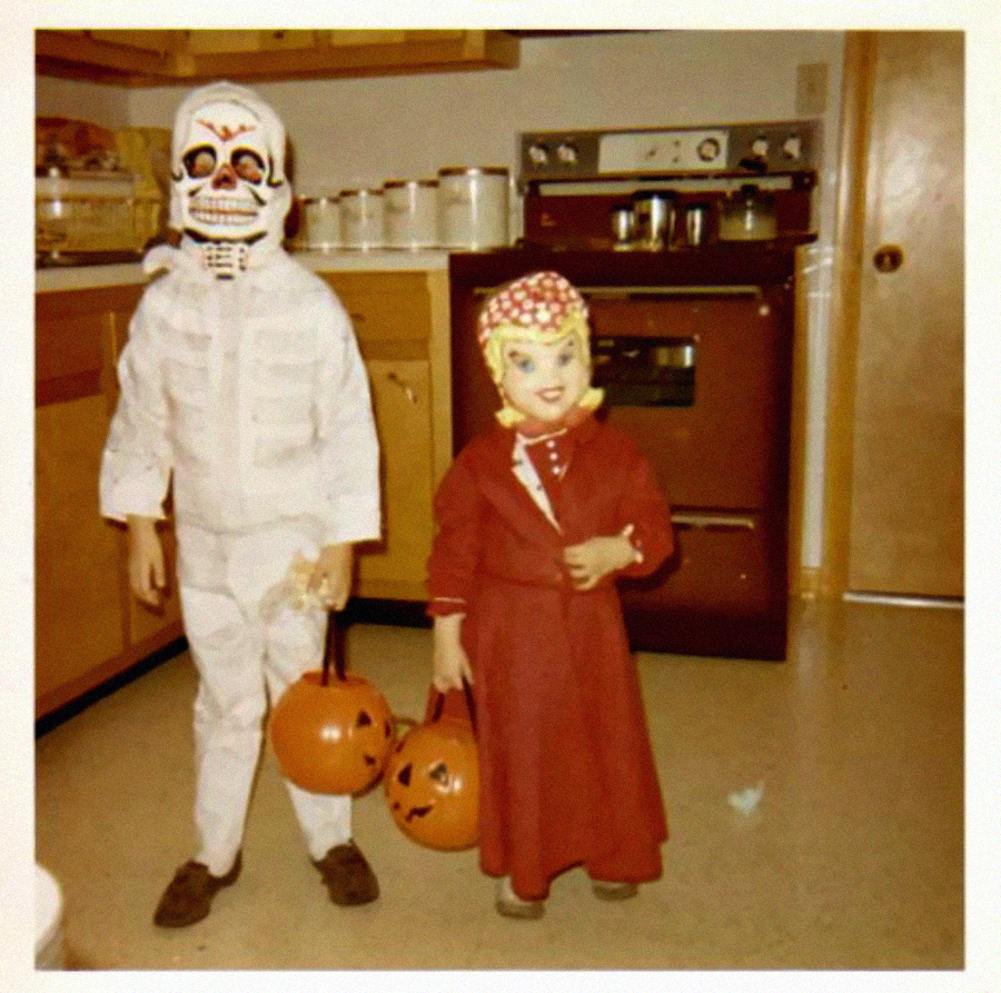 Two children in vintage Halloween costumes stand in a kitchen, each holding a pumpkin-shaped bucket. One wears a skeleton mask and white outfit, the other wears a red dress and a mask with a bonnet.