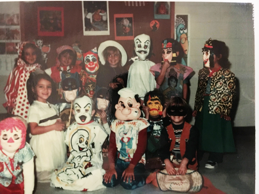 A group of children wearing various Halloween costumes and masks pose together indoors, smiling and sitting or standing in front of a decorated wall.