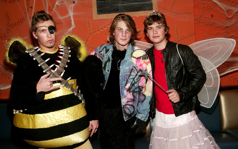 Three people in Halloween costumes: one dressed as a bee with an eyepatch and ammo belt, one in a denim jacket, and one wearing a tiara, fairy wings, and a skirt while holding a wand.
