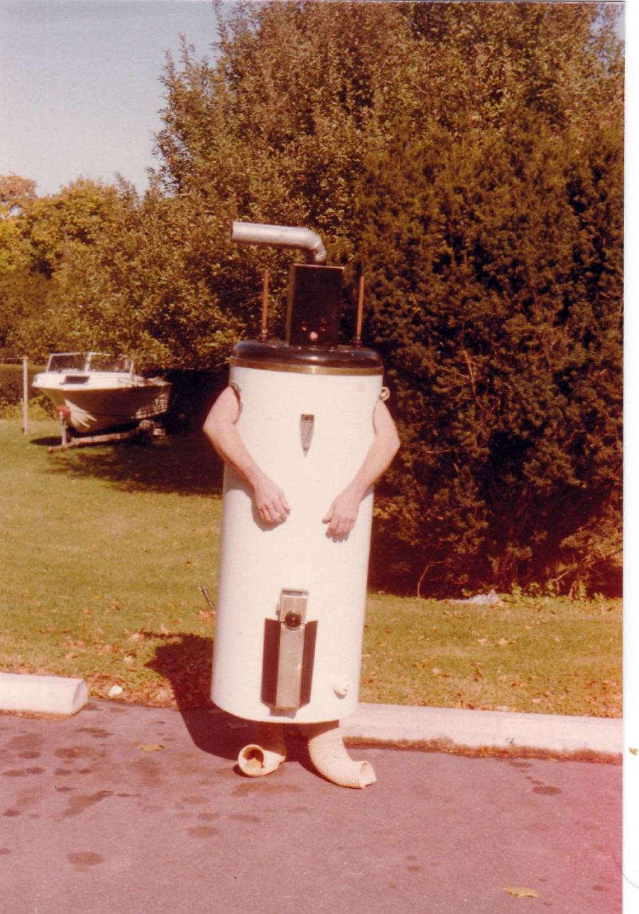 A person stands outdoors dressed in a homemade water heater costume, with their arms and legs visible. Trees and a boat are in the background on a sunny day.