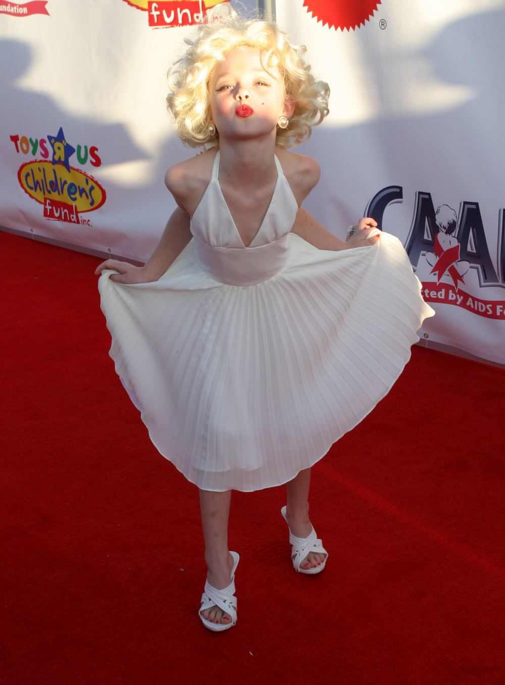 A young girl dressed as Marilyn Monroe in a white halter dress with a pleated skirt, posing on a red carpet.