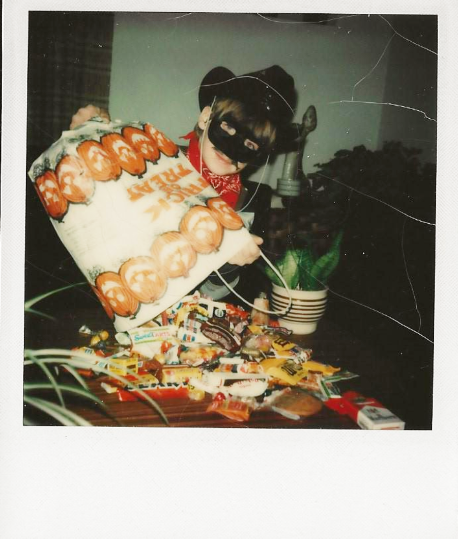A child in a cowboy hat and mask pours Halloween candy from a large bag onto a table. A plant and furniture are visible in the background.