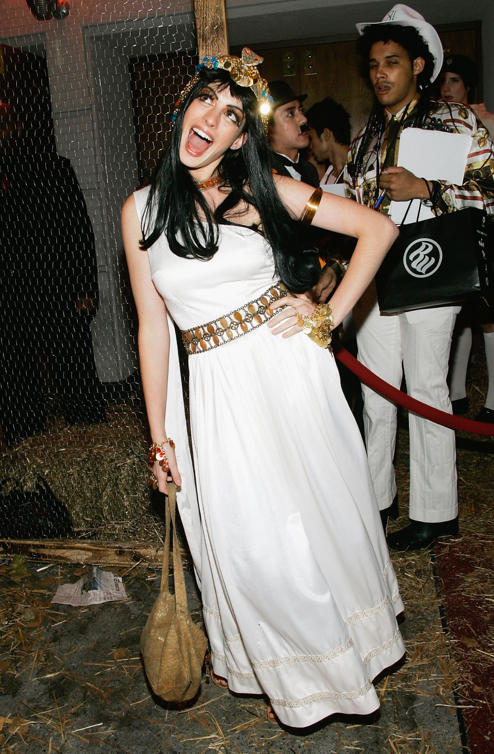 A person in a white Greek-style costume with gold jewelry and a floral headpiece poses with their mouth open.