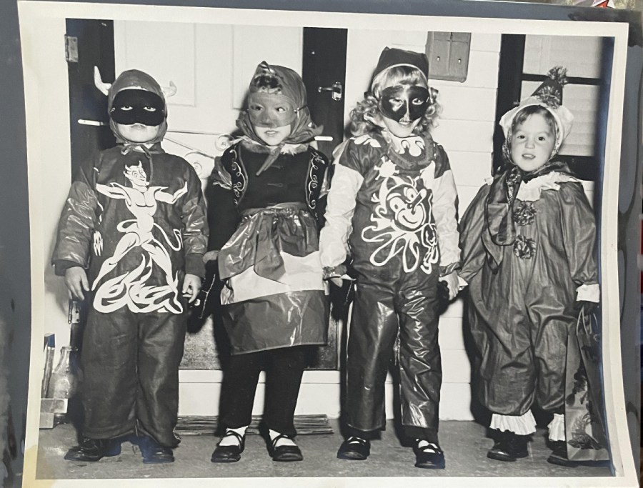 Four children in homemade costumes stand in a row, smiling. Their outfits include masks and decorated clothing, resembling various whimsical or fantasy characters, in front of a house porch. The photo is black and white.