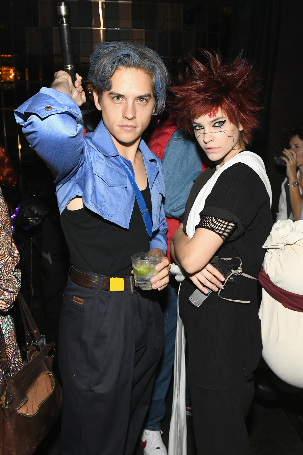 Two people in cosplay costumes: a man in a blue cropped jacket holding a lightsaber handle, and a person with spiky red hair and facial markings posing with arms crossed.