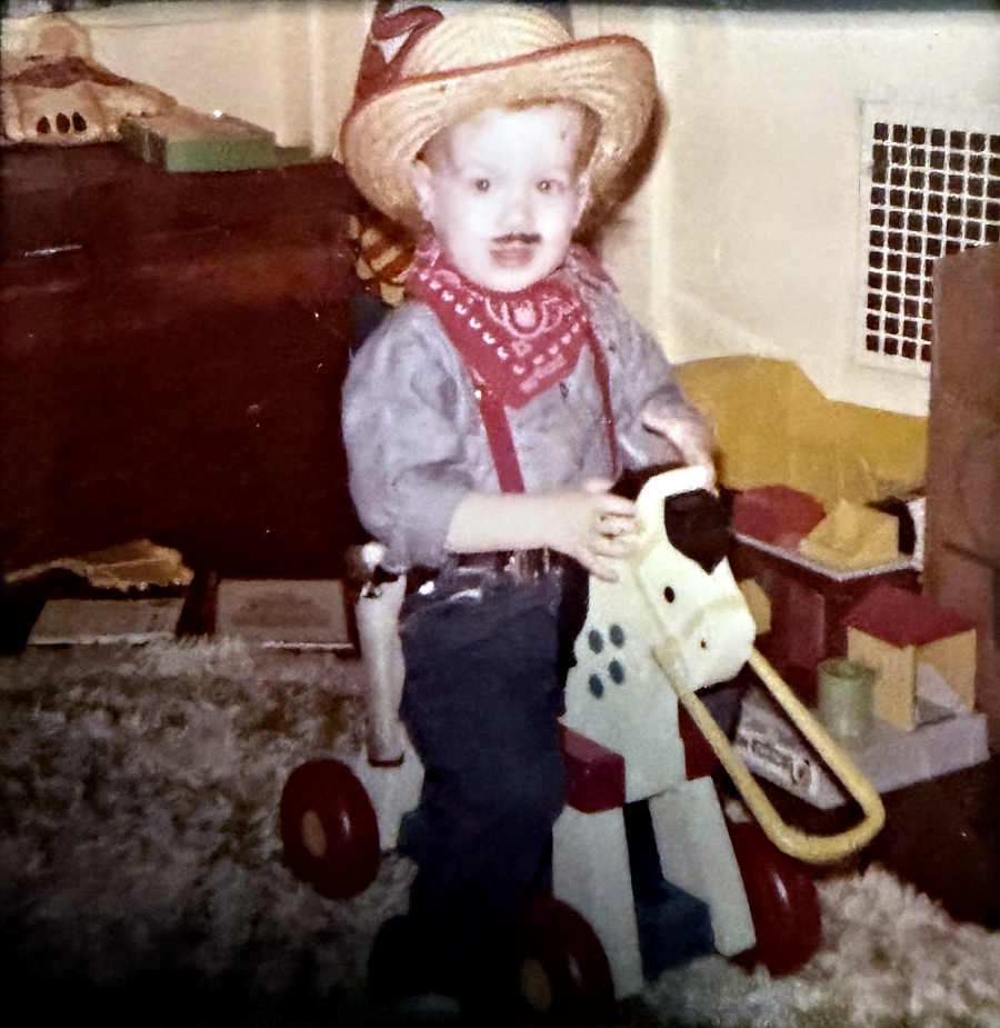 A young child dressed as a cowboy, with a hat, bandana, and suspenders, rides a toy horse on wheels in a cozy room with toys and books scattered around.