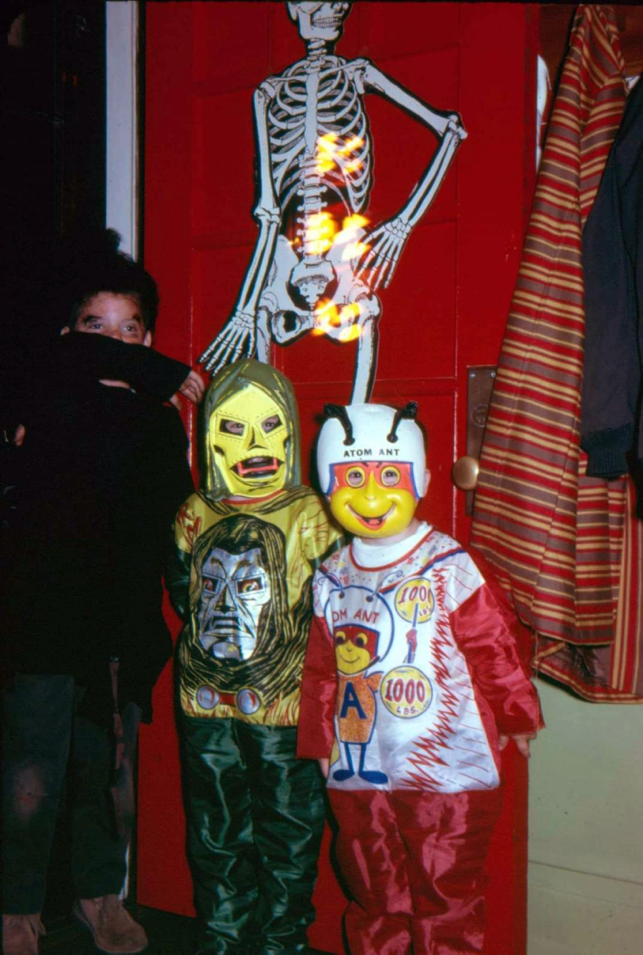 Three children in Halloween costumes stand in front of a red door with a skeleton decoration. Two wear colorful masks and suits, while one stands to the left, partially obscured by a black sleeve covering their mouth.