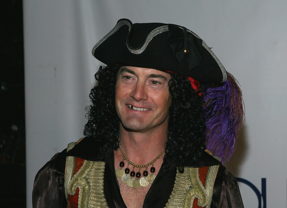 A man wearing a pirate costume with a tricorne hat, a curly black wig, and a gold coin necklace.