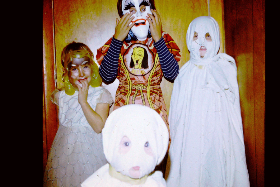 Four children in Halloween costumes: one in a blonde wig, one in a painted mask and robe, one in a ghost sheet, and one in a white hooded outfit, all posing indoors against a wood-paneled wall.