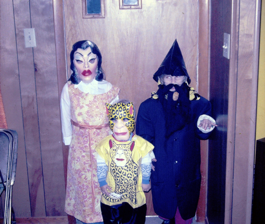 Three children in Halloween costumes stand indoors. One wears a woman’s mask and dress, another is dressed as a leopard, and the third wears a wizard hat and beard. They pose in front of a wood-paneled door.