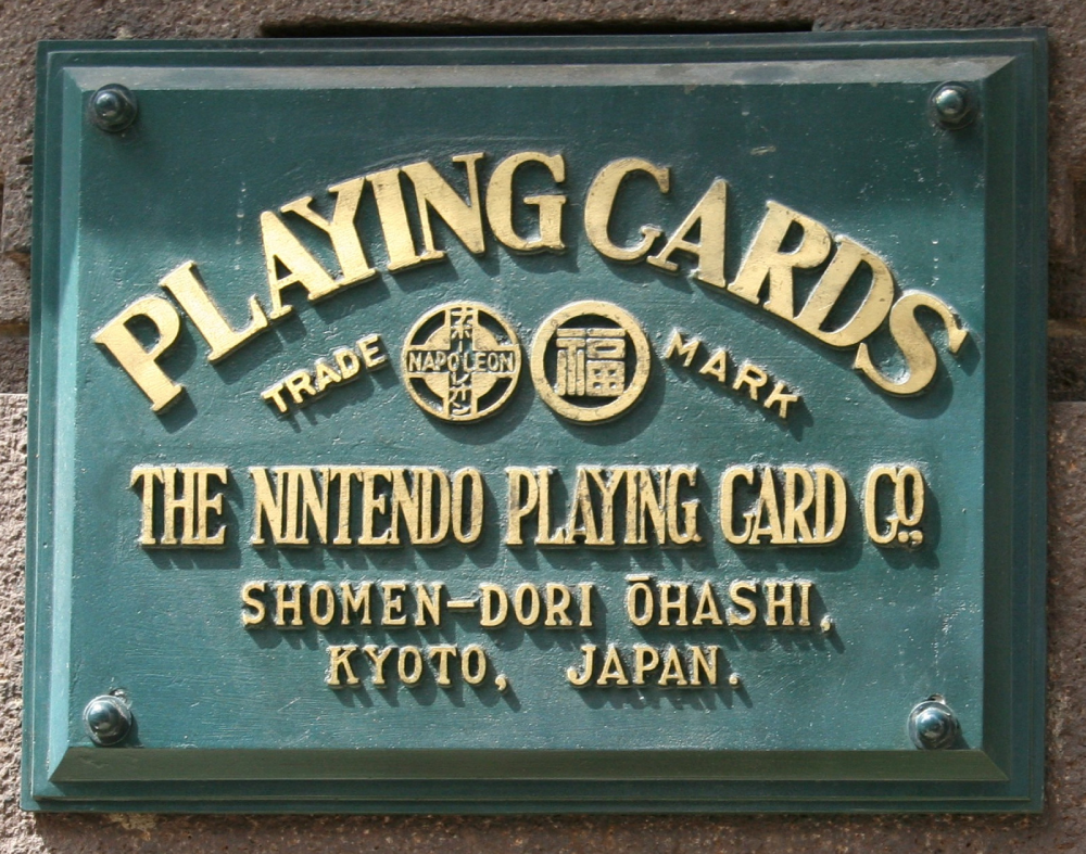 A green plaque with gold text reading: “PLAYING CARDS,” “THE NINTENDO PLAYING CARD CO.,” and the address “SHOMEN-DORI OHASHI, KYOTO, JAPAN.” Includes round logos and the words “TRADE MARK NAPOLEON.”