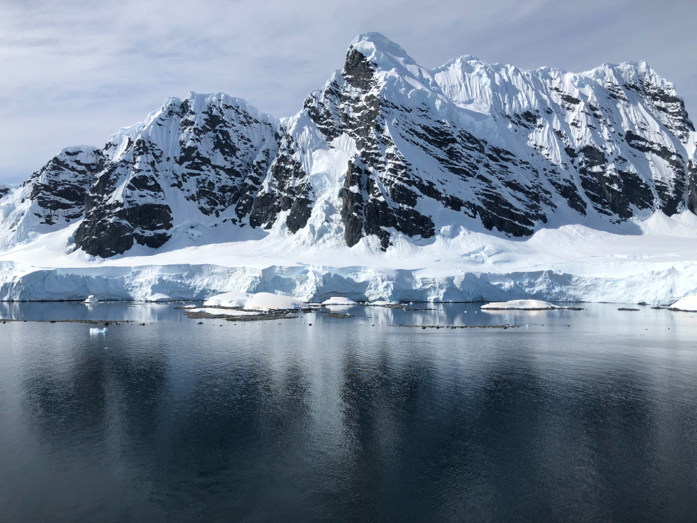 Snow-covered mountains rise above dark, calm water, reflecting the icy cliffs. Patches of blue sky are visible above the rugged, glacier-covered peaks in a pristine polar landscape.