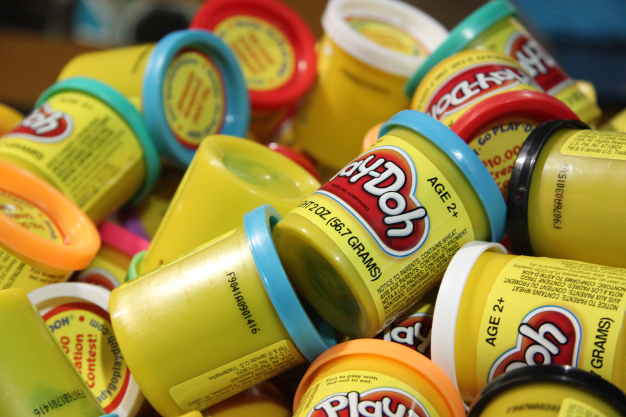 A pile of colorful Play-Doh containers, mostly yellow with different colored lids, some stacked and some lying on their sides. Labels show “Play-Doh” and age recommendations.