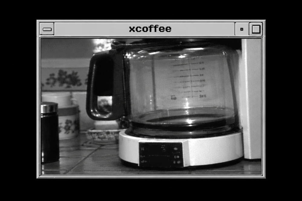 Black and white image shows a nearly empty coffee pot on a coffee maker, viewed through a computer window labeled “xcoffee.” Kitchen items, like jars, are visible in the background.