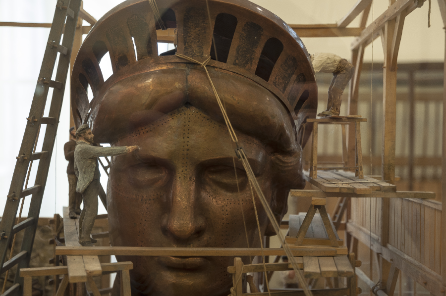 Model workers assembling the head of the Statue of Liberty surrounded by scaffolding, ladders, and wooden platforms in a workshop setting.