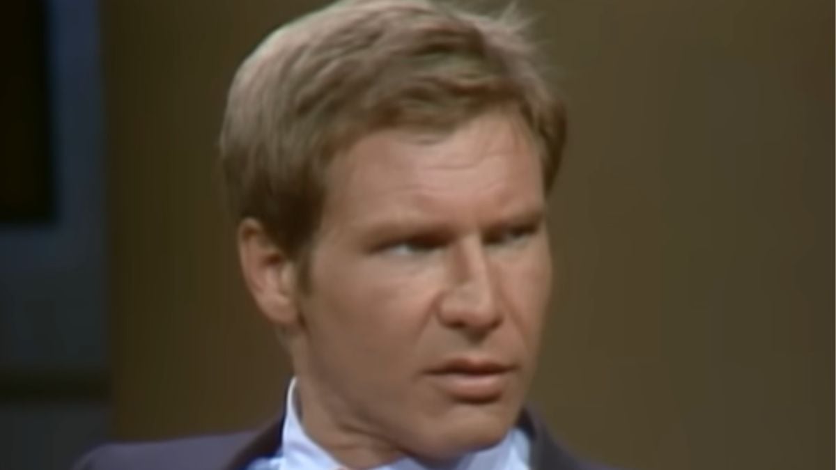 A man with light brown hair wearing a suit and tie looks to the side with a serious expression against a plain, blurred background.