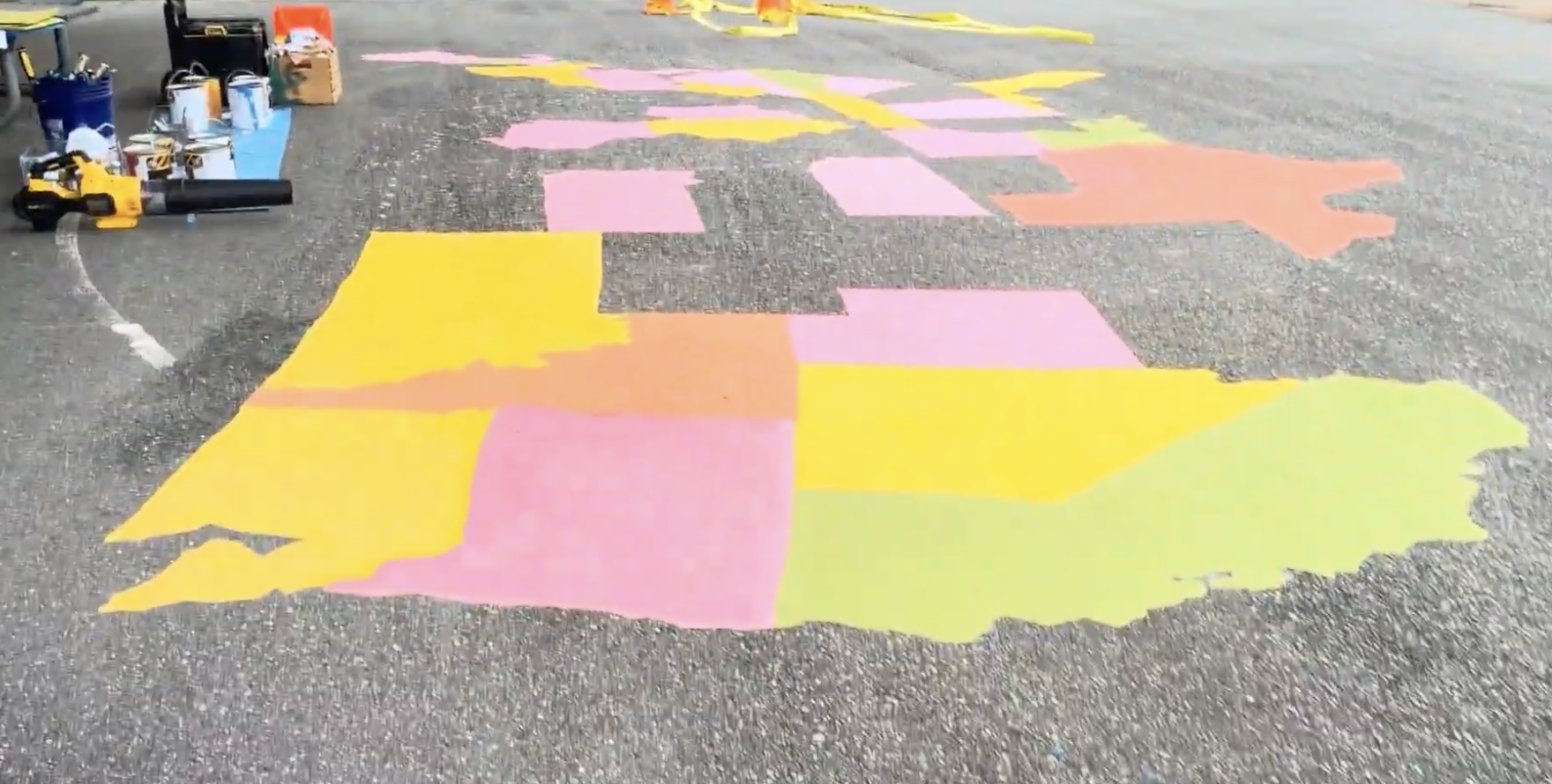 A large map of the United States painted in colorful blocks on an asphalt surface, with painting supplies nearby.