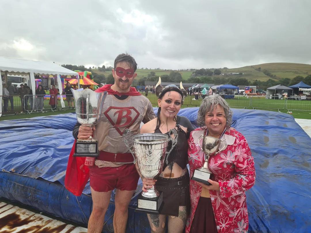 Three people in costumes holding trophies while standing on a large blue inflatable mat at an outdoor event.