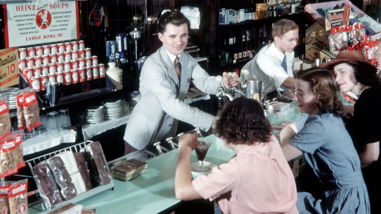 A man in a suit serves ice cream sodas at a vintage soda fountain counter to three women seated on stools, while another worker prepares drinks in the background.