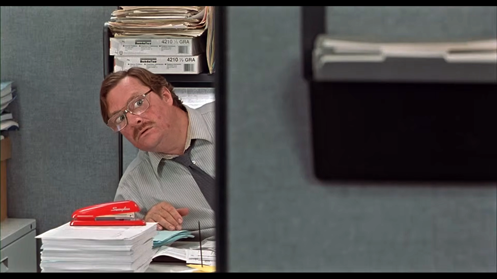 Milton from Office Space peering around a cubicle wall at his desk with a red stapler.