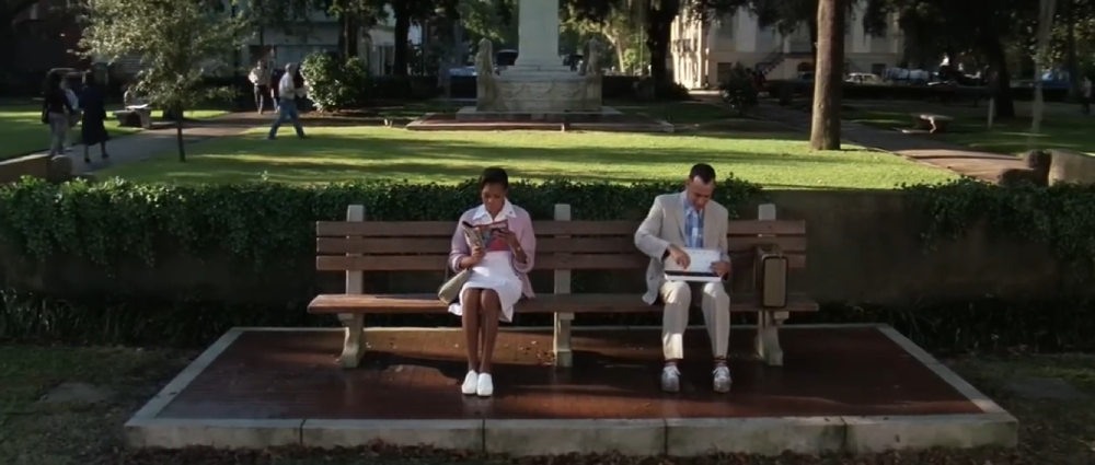 A woman in a white uniform reading a magazine and a man in a tan suit sitting on a park bench.