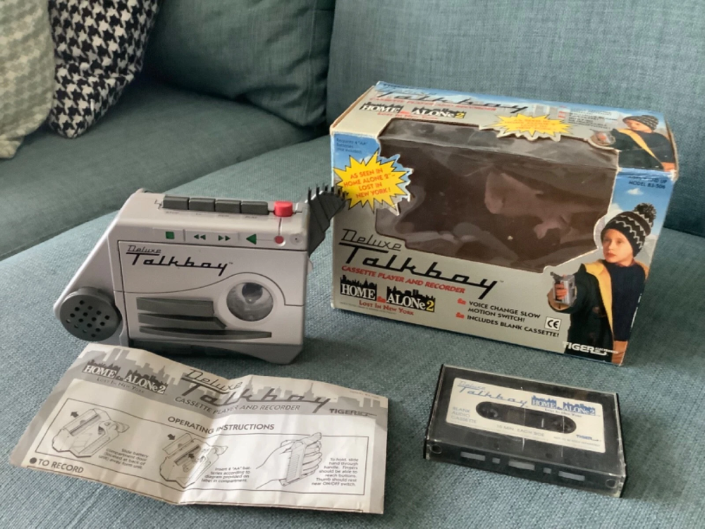 A Deluxe Talkboy cassette player, its original packaging featuring Home Alone 2 branding, an instruction manual, and a blank cassette tape.