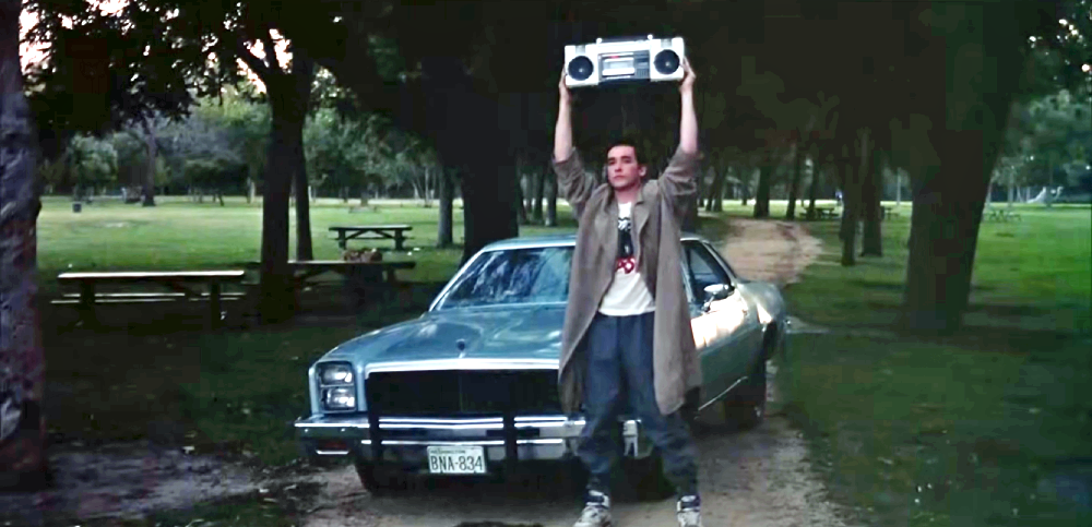 A young man stands in front of a car in a park, holding a large boombox high above his head with both hands.