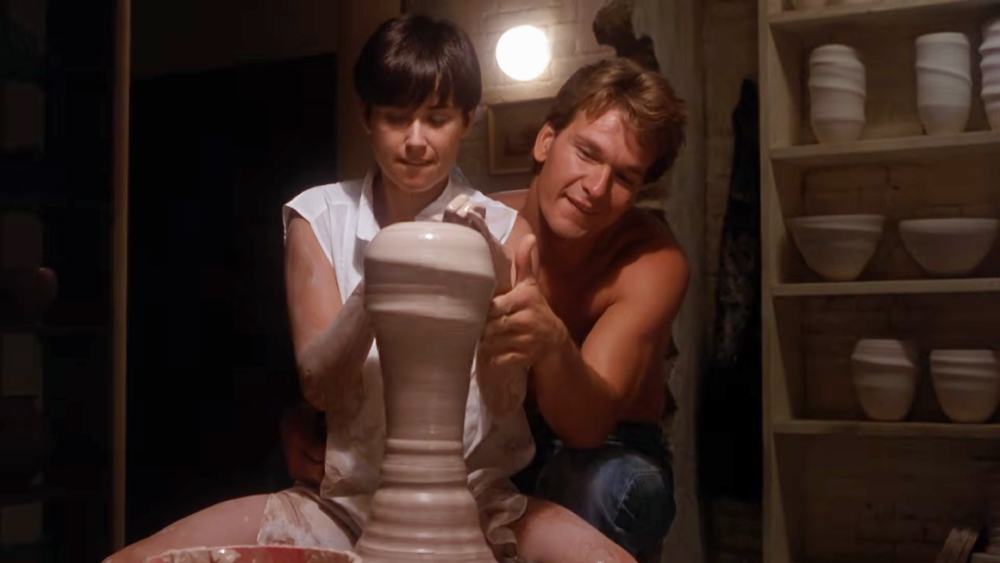 A woman and a man sit together at a pottery wheel, both with their hands on a shaping clay vase.
