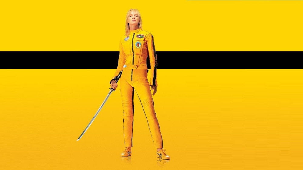 A woman standing against a solid yellow background with a black horizontal stripe, wearing a yellow motorcycle suit and holding a katana.