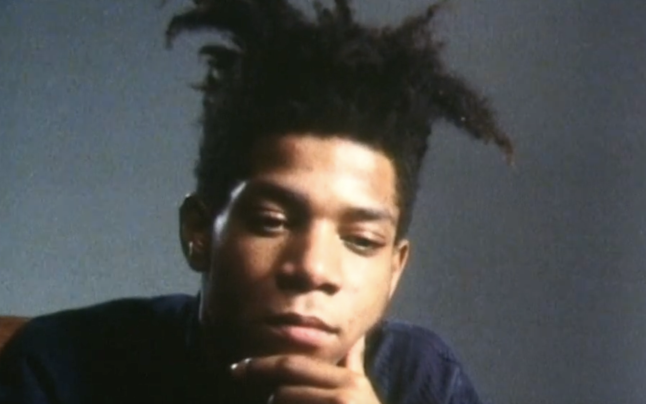 A close-up portrait of Jean-Michel Basquiat looking downward with his hand resting against his chin.