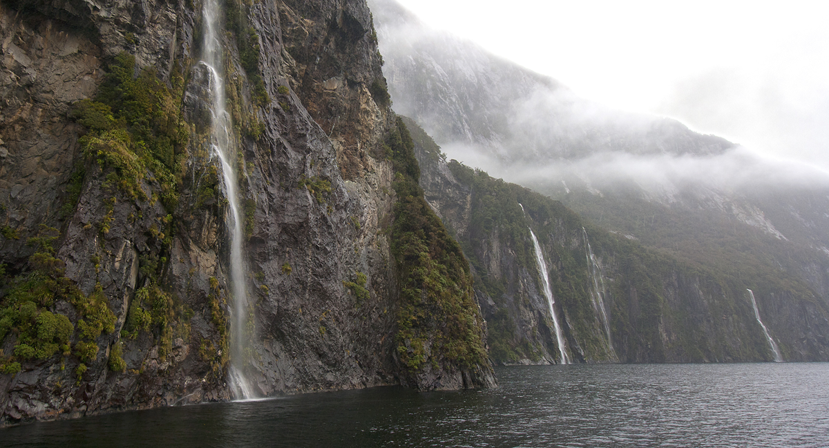 Places in the rain. Multiple waterfalls cascade down steep, mist-covered cliffs into a dark body of water.
