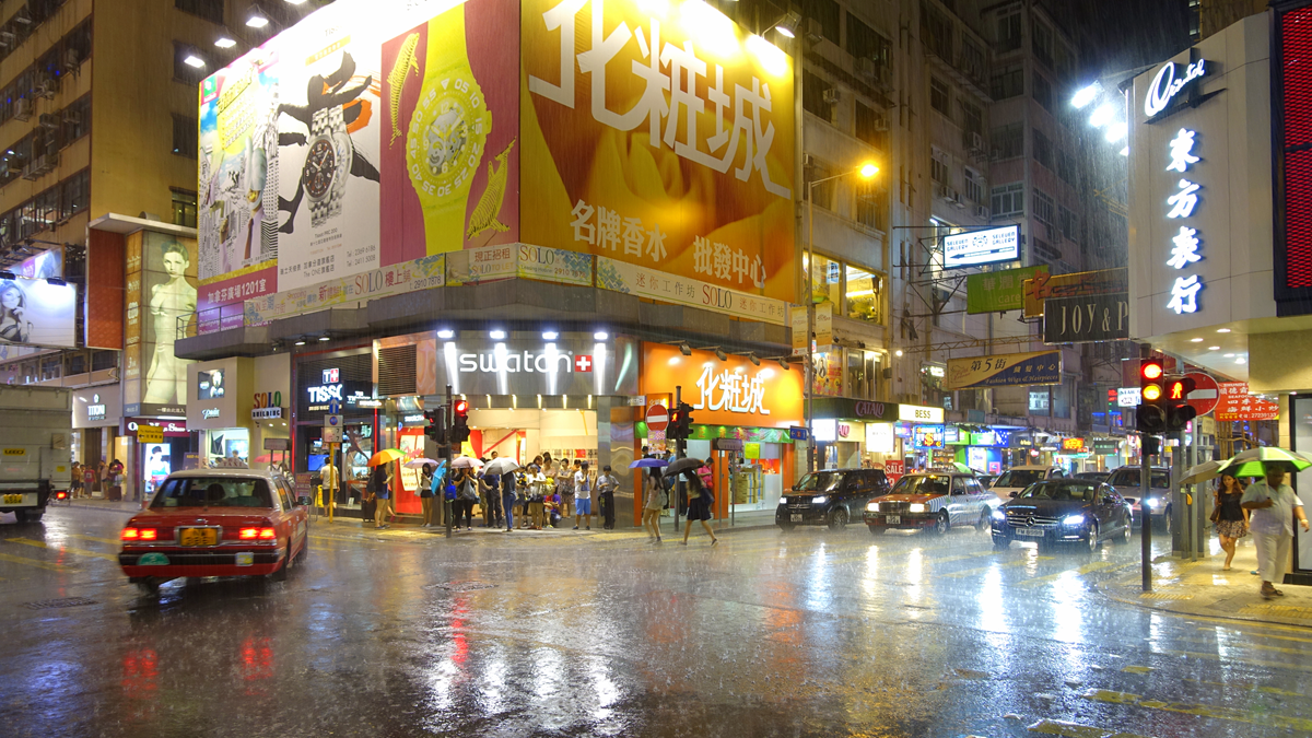 A rainy night street intersection in Hong Kong featuring a brightly lit Swatch store corner, multiple storefront signs with Chinese text, and cars driving on a wet road.