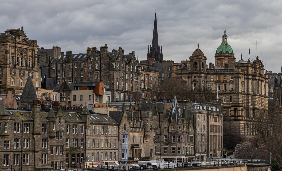 A dense cityscape of traditional stone buildings in Edinburgh, featuring a tall church spire and a prominent dome under a cloudy sky.