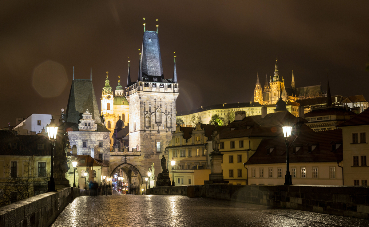 The Charles Bridge in Prague at night, featuring a stone tower and illuminated historical buildings in the background.