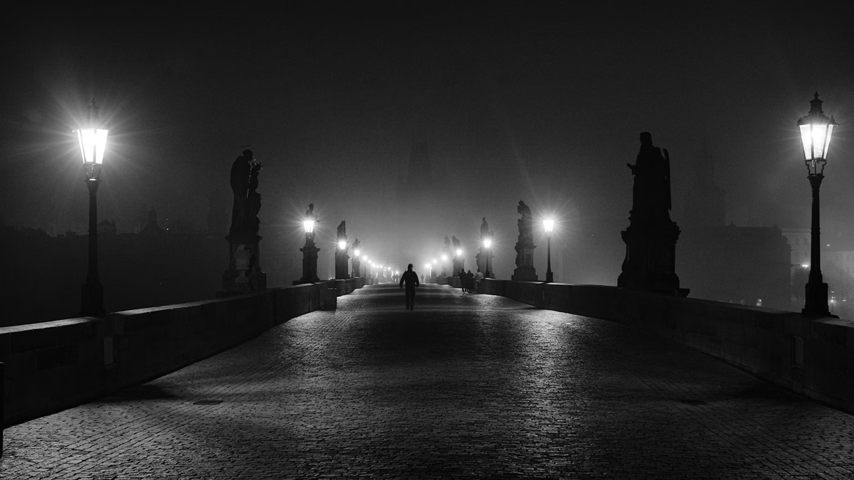 A lone person walks across a fog-covered stone bridge lined with glowing street lamps and statues at night.