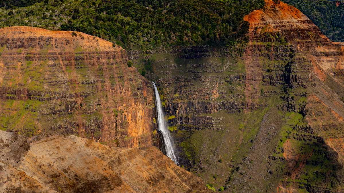 A slender waterfall cascades down a steep, rocky canyon wall with green vegetation and patches of red earth.