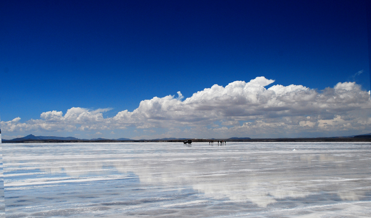 A small group of people standing on the Salar de Uyuni salt flat under a vast blue sky with reflective cloud cover.