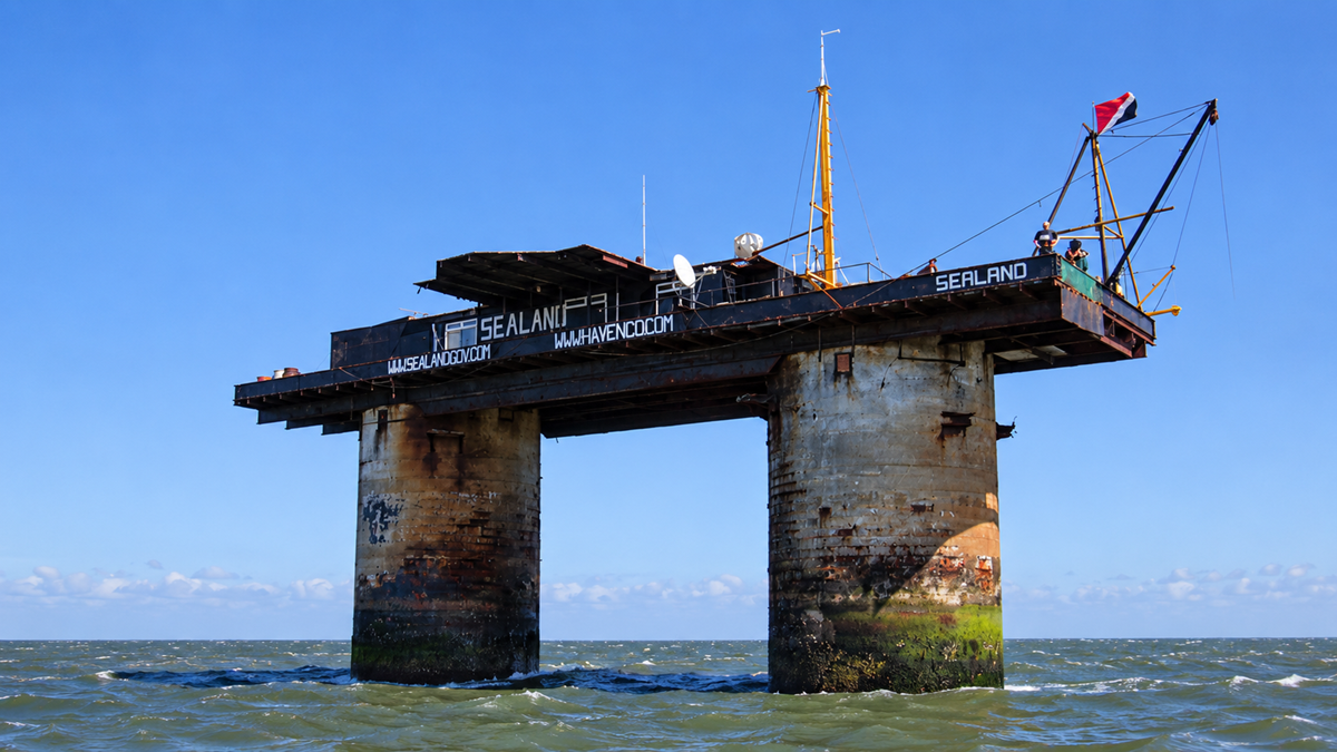 Micronations. A sea fort with two concrete pillars and a platform above the waves, featuring antennas, rust, and a red flag, marked “Sealand.” The sky is clear and blue, and the water is choppy.