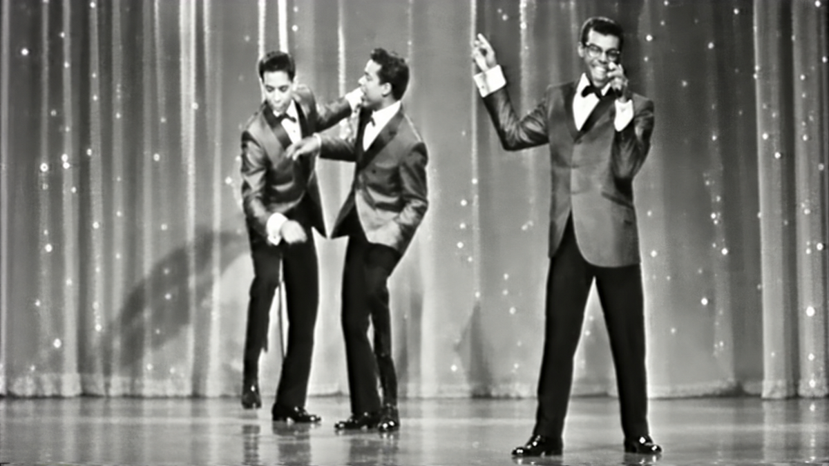 Three men in tuxedos performing on a stage with a shimmering curtain backdrop.