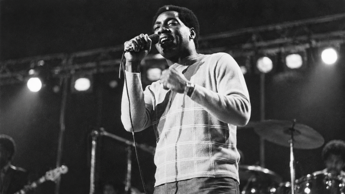 Black and white photo of a man singing into a microphone on stage with a band.