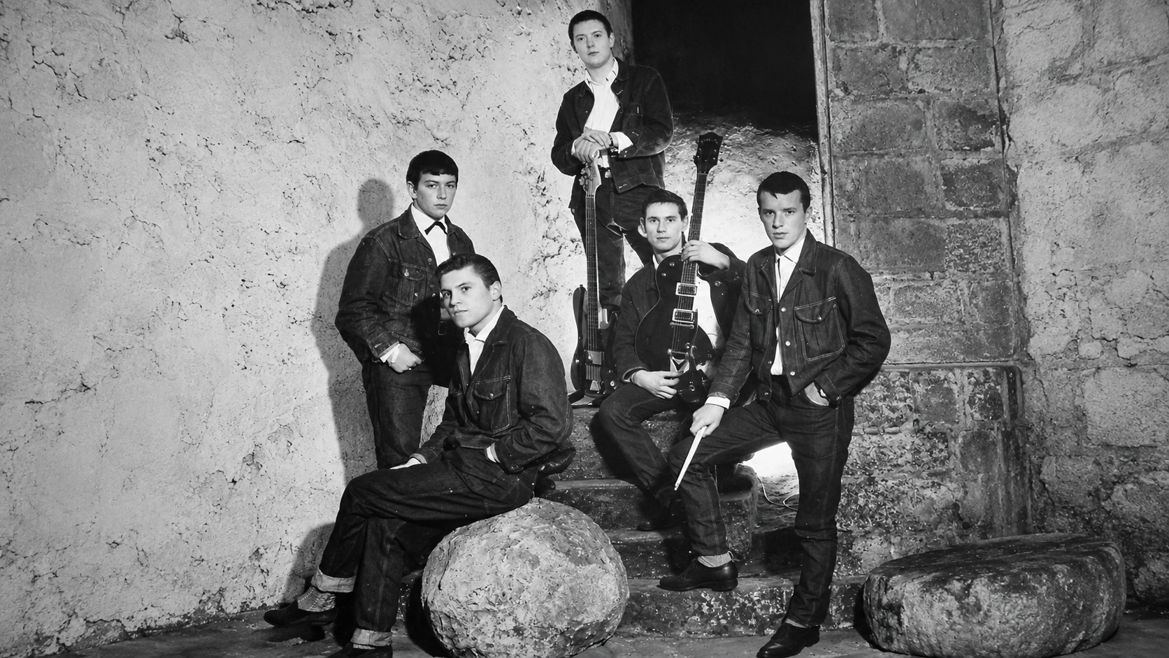 A black-and-white group portrait of five young men dressed in denim jackets, with two posing with guitars, in a stone room.