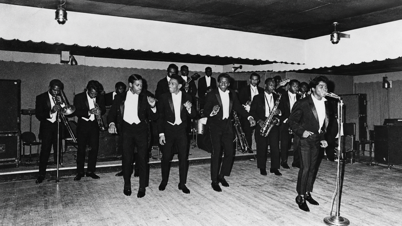 James Brown performing on stage with his band, all wearing tuxedos.