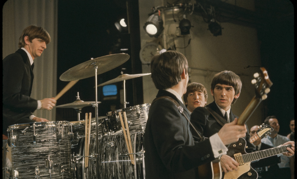 The Beatles performing on stage, with Ringo Starr on drums and Paul McCartney, John Lennon, and George Harrison playing instruments.