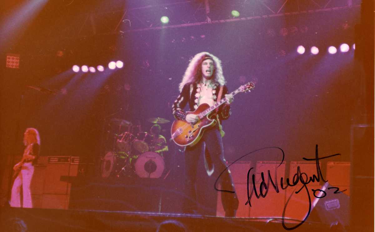 Ted Nugent performing on stage with a guitar, with a large autograph across the lower right.